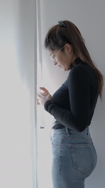 窓の前に立っている若い十代の女の子は 彼女をチェックし 微笑んでいます 自宅で勉強するコロンビアの大学の学生 1080 垂直ビデオ — ストック動画