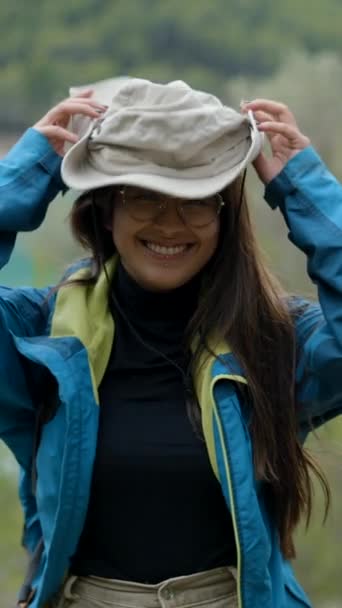 自然の中で若い暗い髪の探検家女性は帽子と笑顔をかける 垂直ビデオ — ストック動画