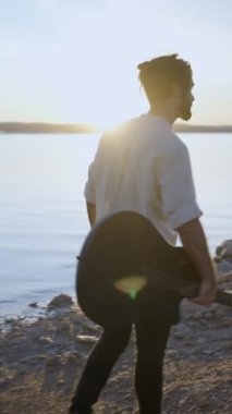 Gitarlı hippi, göl kıyısında güneşin altında gün batımını seyrediyor.