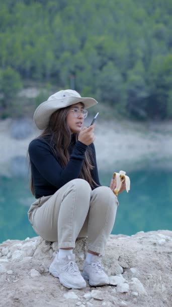 Mädchen Mit Hut Sitzt Auf Stein See Isst Banane Und — Stockvideo