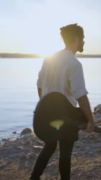 ギターを持つヒップスター男性は 太陽に対してスタニービーチを歩く湖岸で日没を楽しむ スローモーションバックビュー 垂直フルHdビデオ — ストック動画