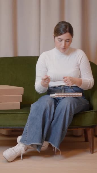 若い白人起業家女性はレトロスタイルのソファーに座っている間ノートにノートを取ります 垂直フルHdビデオ — ストック動画