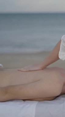 Plajda tedavi masajı yapan beyaz, ikili olmayan genç bir insan. Masör sırt masajına baskı uyguluyor - FullHD dikey video