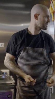 Restoran mutfağında önlük bağlayan kel aşçı FullHD dikey video.