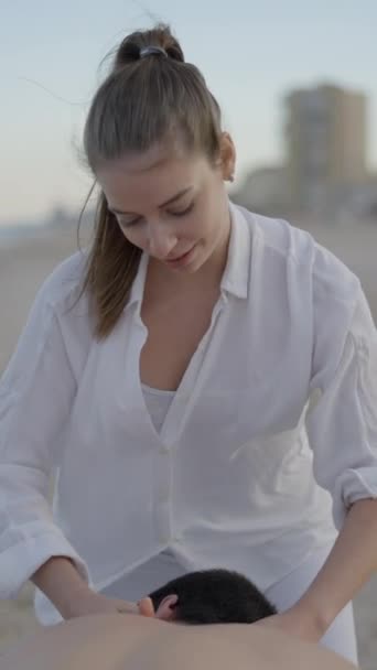 海辺で男性にマッサージをするプロの若い女性 バックグラウンドの明確な空と波 Fullhd垂直ビデオ — ストック動画