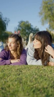 Genç kızlar çim üzerinde yüzükoyun yatar ve konuşurlar, cephe manzarası - FullHD dikey video