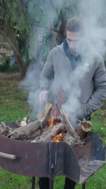 Genç adam BBC 'deki çubukları hareket ettiriyor, yeşil arka bahçede yemek pişirmek için yakacak odun yakıyor.
