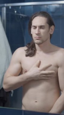 Genç çıplak bir adam banyo aynasının önünde kaslarına ve göğüslerine dokunuyor.