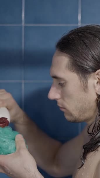シャワーの浴室スポンジにいくつかの浴室石鹸を注ぐ若い髪の男 クローズアップショット 垂直ビデオ — ストック動画