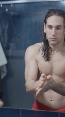 Genç metroseksüel adam duş aldıktan sonra banyo aynasının önünde saçı için serum kullanıyor.