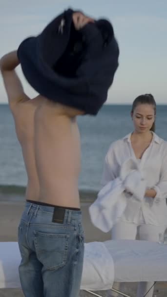 クライアントはビーチに到着し マッサージを受けるためにセーターを離れます 白い服を着た女性マッサージ師 フルHd垂直ビデオ — ストック動画