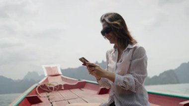 Güneş gözlüklü Asyalı bir kadın cep telefonuna bakar ve gülümser.