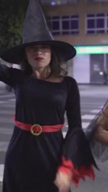 Cadı Cadılar Bayramı kostümlü iki Kolombiyalı kız. Süpürge çubuklarıyla, gece kaldırımda dikilirken gülümseyen kameraya bakıyorlar. - Orta yakın çekim - Dikey 1080