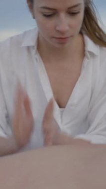 Tanınmayan adam sahilde yağ ile sırt masajı yaptırıyor. Güçlü elleri olan genç beyaz kadın masaj yapıyor - FullHD dikey video