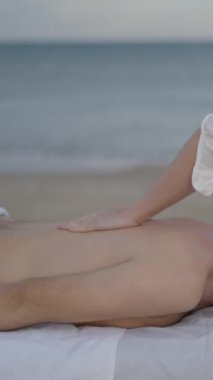 Plajda tedavi masajı yapan beyaz, ikili olmayan genç bir insan. Masör sırt masajına baskı uyguluyor - FullHD dikey video