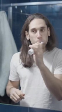 Genç, beyaz bir adam, banyo aynasının önünde dişlerini fırçalıyor.
