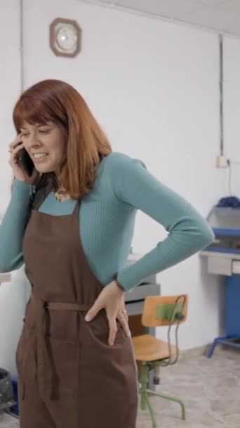 彼女のスマートフォンで話すフリーランスのジュエラーは 彼女のワークルームでエプロンを着用しています 中型ショットフルHd垂直ビデオ — ストック動画