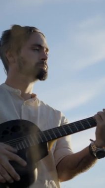Açık havada gitar çalan İspanyol sakallı adam güneş ışığıyla aydınlatılan mavi gökyüzüne karşı - Dikey FullHD video