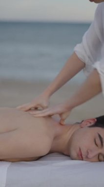 Kumsalda rahatlatıcı masaj seansında beyaz bir erkeğe yaklaş. Erkek uzanır ve masaj yaptırır - FullHD dikey video