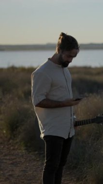 Gitarist bir gitarist gün batımında nehir kenarında dikey FullHD videosu çekerken cep telefonuyla arama yapıyor.
