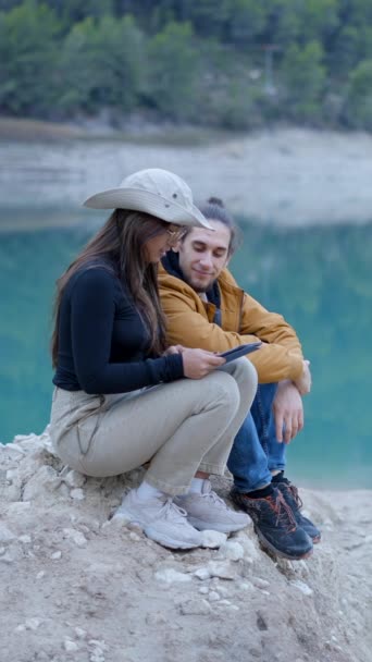 快乐的年轻夫妇坐在岩石上 在静谧的湖畔和森林中使用石碑 垂直视频 — 图库视频影像