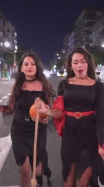 Cadı kostümlü iki güzel Kolombiyalı kız, biri elinde balkabağı, Valencia, İspanya 'da gece sokakta yürürken eğleniyor. - Tam Vuruş - Dikey 1080