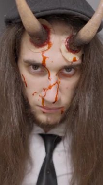 Alnında plastik boynuzları ve yüzünde sahte kanı olan uzun saçlı beyaz adam ciddi ciddi bakıyor - 1080 dikey video