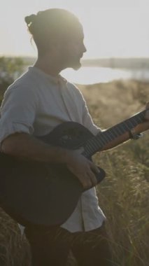 İspanyol erkek gitarist Sihouette gün batımında göl kenarında çimenli bir çayırda akor çalıyor ve şarkı söylüyor.