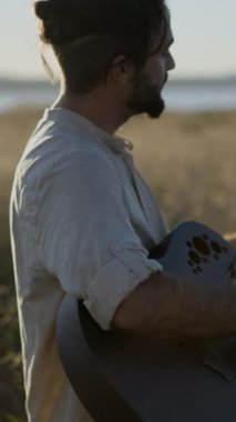 Man Gitarist İspanya 'da çimenli bir alanda duruyor ve güzel güneş ışığına karşı siyah akustik gitarla akorlar çalıyor - yavaş çekim profil görüntüsü - Dikey FullHD video