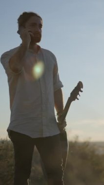 Genç sakallı gitaristin portresi, günlük beyaz gömlekli cep telefonuyla konuşan, elinde gitar tutan açık havada gölün üzerinde gün doğumuna bakan dikey FullHD videosu.