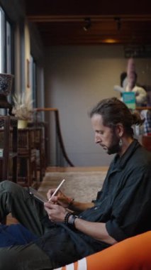 Sakallı bohem adam tabletini etnik açıdan dekore edilmiş FHD dikey video odasında çalıyor.