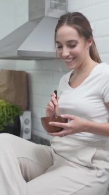 Mutfak tezgahında oturan aktif kadın. Tahta bir kasede yemek yerken dans etmek. orta çekim - Dikey