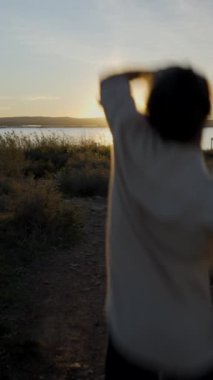 Bir gitarla atlayan adamın gölün üzerinden gün batımına bakışı - Dikey FullHD videosu