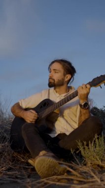 Çimenli bir çayırda oturan ve günbatımında gitar çalan genç bir adam - yavaş çekim - Dikey FullHD video