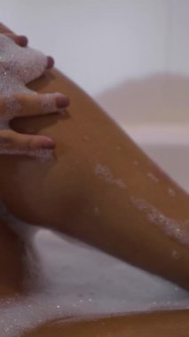 Kadın elleri köpük dolu bir küvette bacaklarını okşar - FHD dikey video
