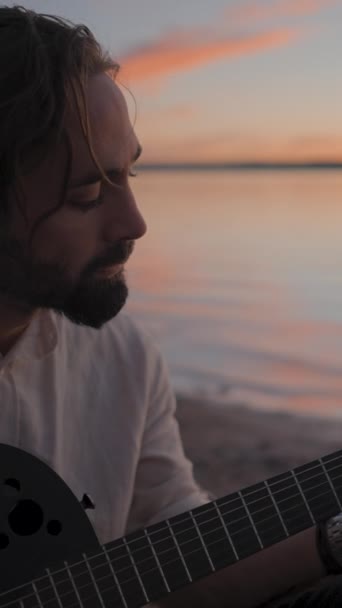 在湖畔对着夕阳的天空弹奏声吉它的多愁善感的嬉皮士人 慢镜头 垂直Fullhd视频 — 图库视频影像