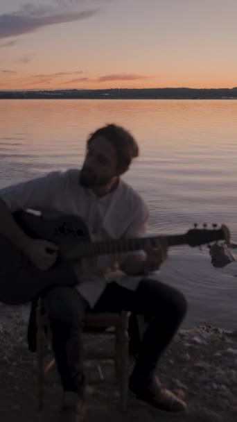 西班牙落日下 一位吉他手在湖边的声学吉它上弹奏和弦的轮廓 Gimbal拉回 垂直Fullhd视频 — 图库视频影像