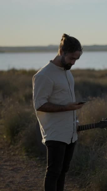 ギターを持っている男ギタリストと日没の川岸に立っている間携帯電話を使用して通話をする 垂直フルHdビデオ — ストック動画