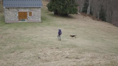 Kafkasyalı, şapkalı ve sırt çantalı yetişkin bir kadın köpeğiyle dağda yürüyor. 4K yatay video.