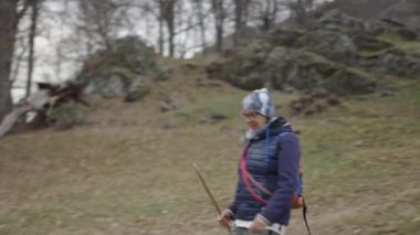 Sırtında bir sırt çantası ve köpeğiyle birlikte bir baston ve sıcak kıyafetle dağlarda yürürken olgun bir kadının ağır çekimleri - 4K Yatay Video