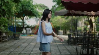 Yavaş çekim genç Asyalı kız elinde akıllı telefonuyla bahçenin yanındaki barın terasında el sallıyor ve gülümsüyor. 4K Yatay video.