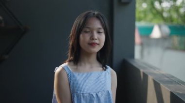 Yavaş çekim genç Asyalı kız kameraya bakıyor. Yazın terasın yanında bir elbise giyip gülümsüyor. 4K yatay video.