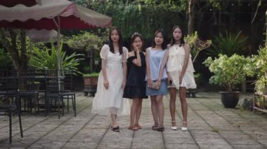 Ağır çekimde dört genç Asyalı kız şık yaz kıyafetleri içinde ellerinde akıllı telefonlarıyla bahçenin yanında 4K yatay video.