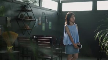 Yavaş çekim genç Asyalı kız sakince yürüyor ve elinde akıllı telefonuyla yazın barın terasında gülümsüyor - 4K yatay video.