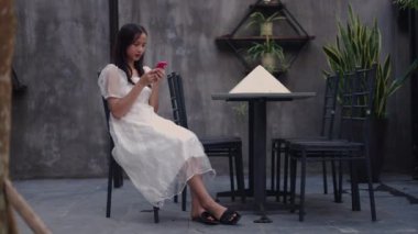 Genç Asyalı kız teras sandalyesinde oturuyor. Yazın akıllı telefonuna danışıyor. 4K yatay video.