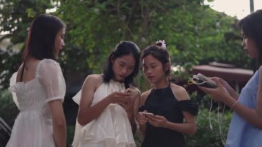 Dört genç Asyalı genç kız akıllı telefonlarına bakarak daire oluşturuyorlar ve yazın bahçedeki fotoğraflar hakkında yorum yapıyorlar - 4K Yatay Video