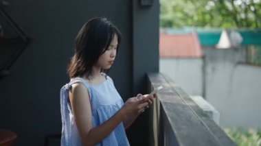 Ağır çekimde genç Asyalı kız ahşap terasta akıllı telefonuna danışmanlık yapıyor. Kameraya mutlu bir şekilde bakıyor. 4K yatay video.