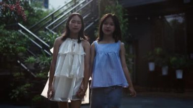 Ağır çekimde iki genç Asyalı kız el ele tutuşup kameraya doğru yürüyorlar yazın bahçede güneşin altında mutlu mesut yürüyorlar - 4K yatay video.