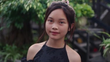 Asyalı genç kızın yüzüne yavaşça zum yap. Bahçede mutlu bir şekilde kameraya gülümse. 4K yatay video.