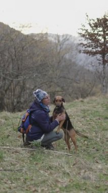 Sırtında şapka ve sırt çantasıyla beyaz, olgun bir kadın Alman çoban köpeği FullHD dikey video köpeğini okşuyor.